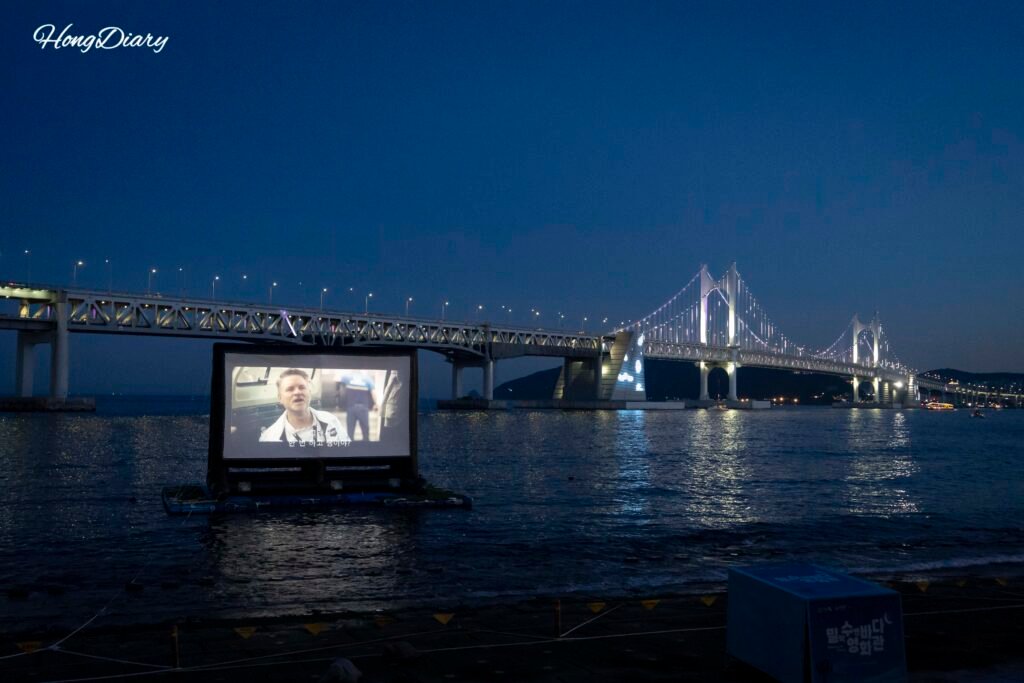 Outdoor movie screen at Millak Ocean Cinema with Gwangan Bridge visible in the background at dusk