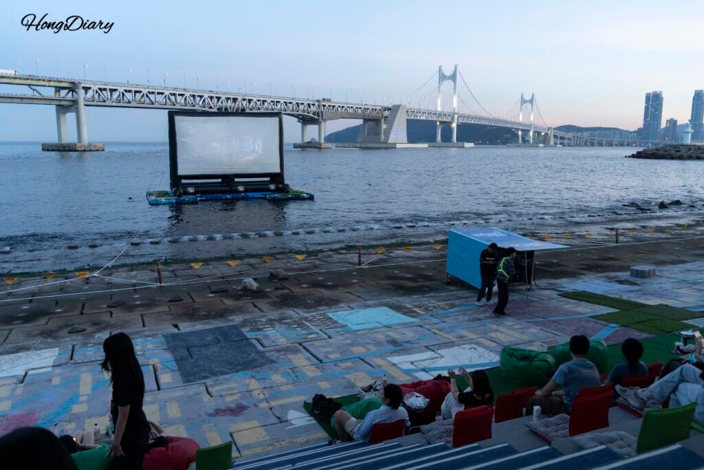 Outdoor movie screen at Millak Ocean Cinema with Gwangan Bridge visible in the background at dusk