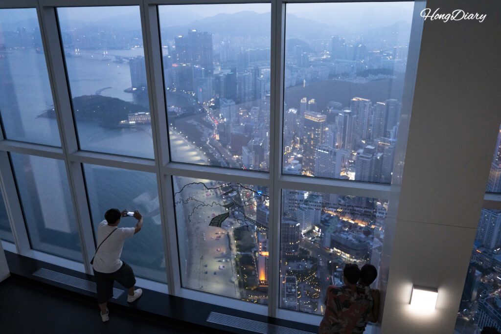 Visitantes fotografiando el paisaje urbano de Haeundae al anochecer desde el interior de Busan X the Sky