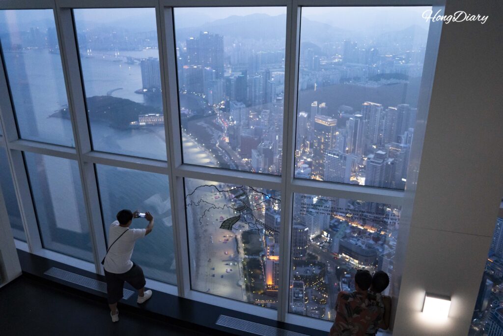 Visitors photographing the early evening Haeundae cityscape from inside Busan X the Sky observatory