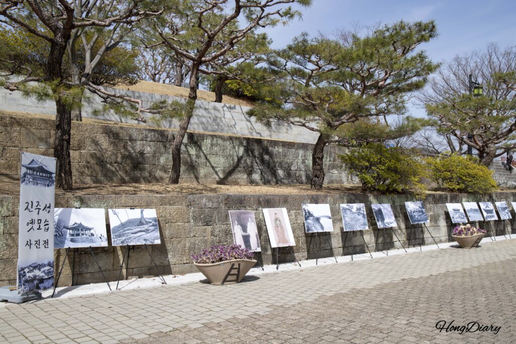 Outdoor photo exhibition displaying historical images of Jinjuseong Fortress along a stone wall path