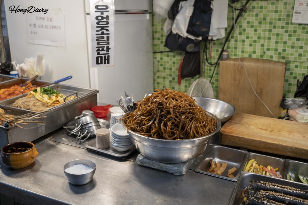 Puesto de kimbap de bardana en el Mercado Seongdong de Gyeongju con montañas de raíz de bardana guisada junto a los rollos recién hechos