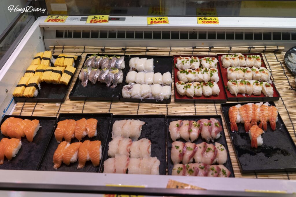 Display case at a Karato Market sushi stall showing egg sushi, salmon, and flounder nigiri priced between 100 and 150 yen per piece