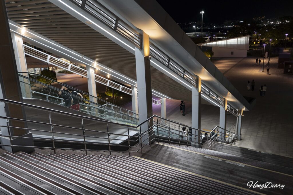Visitantes bajando por escaleras y escalera mecánica desde el puente peatonal hacia North Port Waterfront Park en Busan