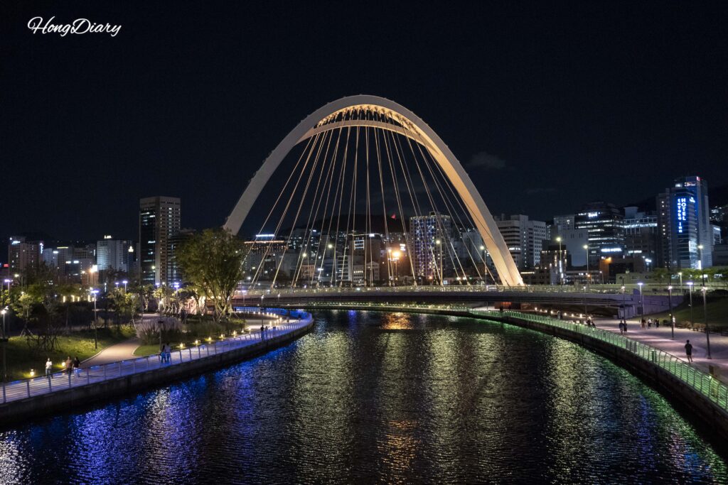 Sendero iluminado y puente peatonal con luz nocturna en North Port Waterfront Park, visitantes caminando