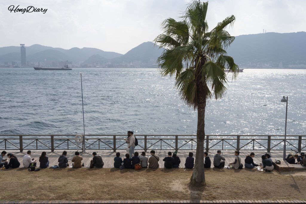 Visitors eating sushi outdoors at Karato Market with the Kanmon Strait and Mojiko visible across the water