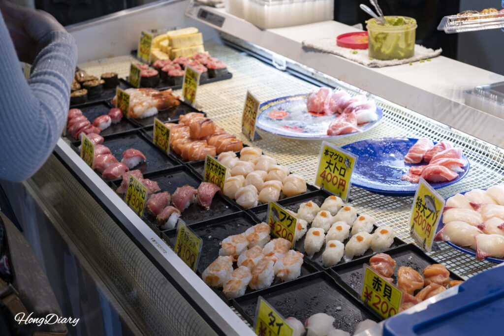 Tourists selecting sushi pieces from a refrigerated display case at Karato Market in Shimonoseki