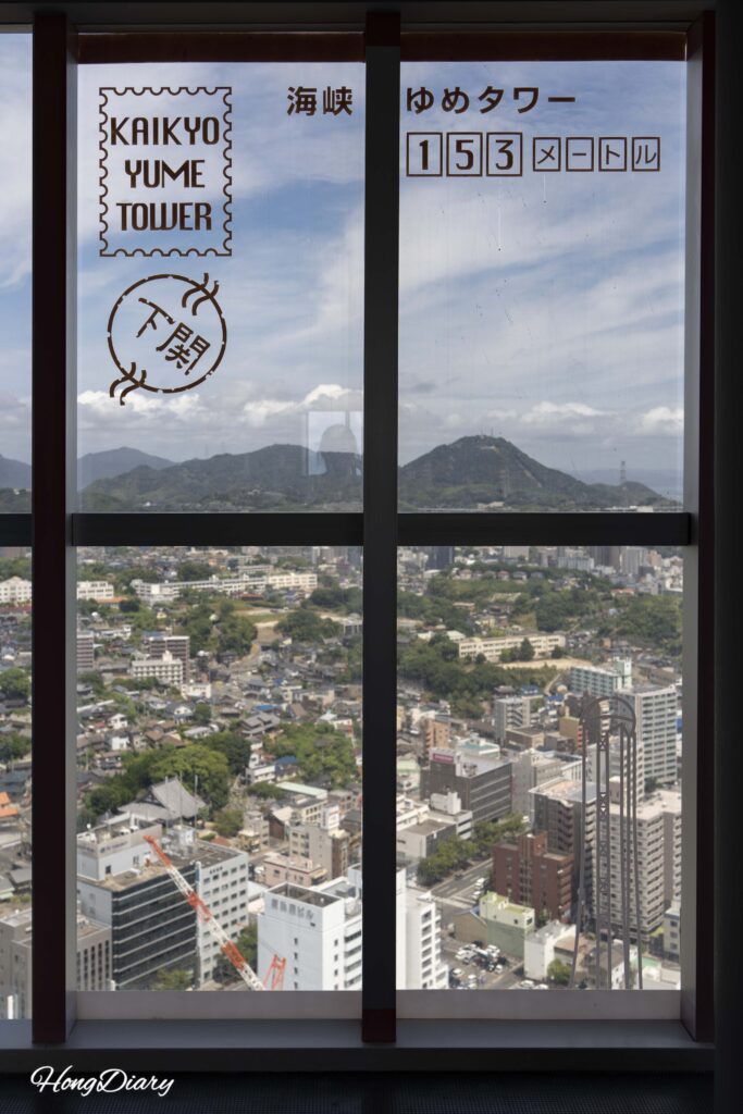 Vista desde el piso 28 de Kaikyo Yume Tower a través de un cristal semitranslúcido con el nombre de la torre y el código postal impresos, creando un efecto visual tipo postal