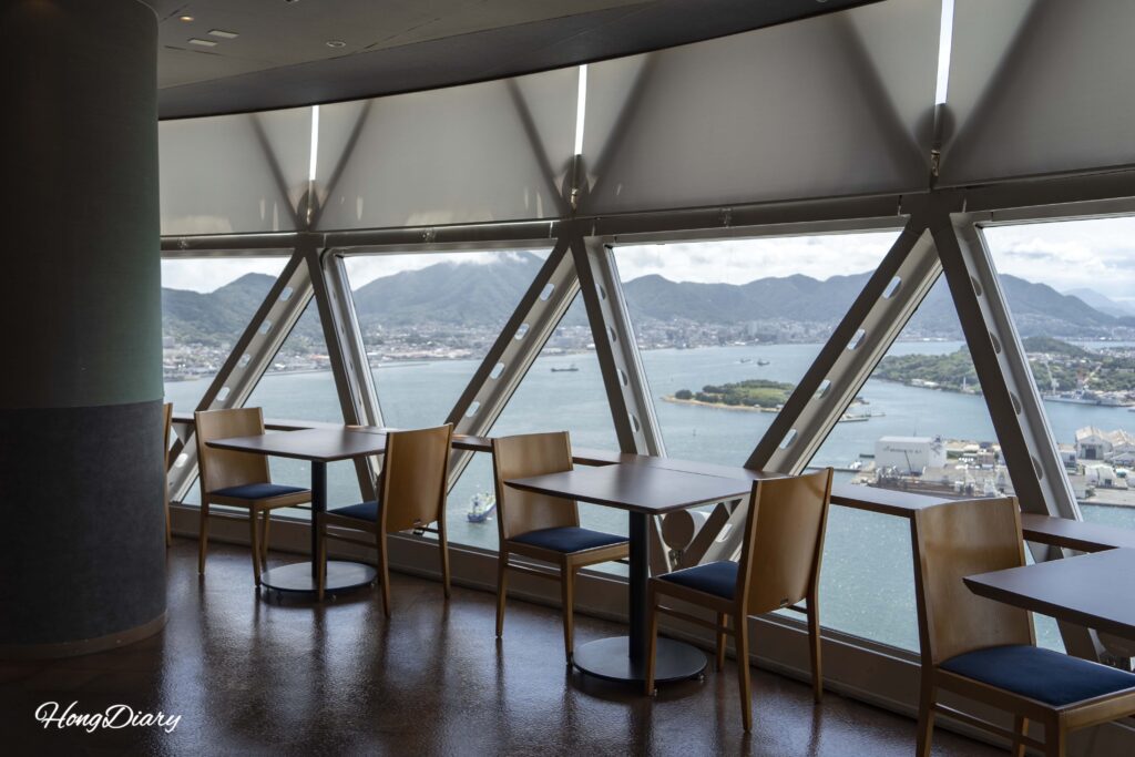 Sala de descanso en el piso 29 de Kaikyo Yume Tower con mesas y sillas de madera y vista hacia el norte del centro de la ciudad de Shimonoseki