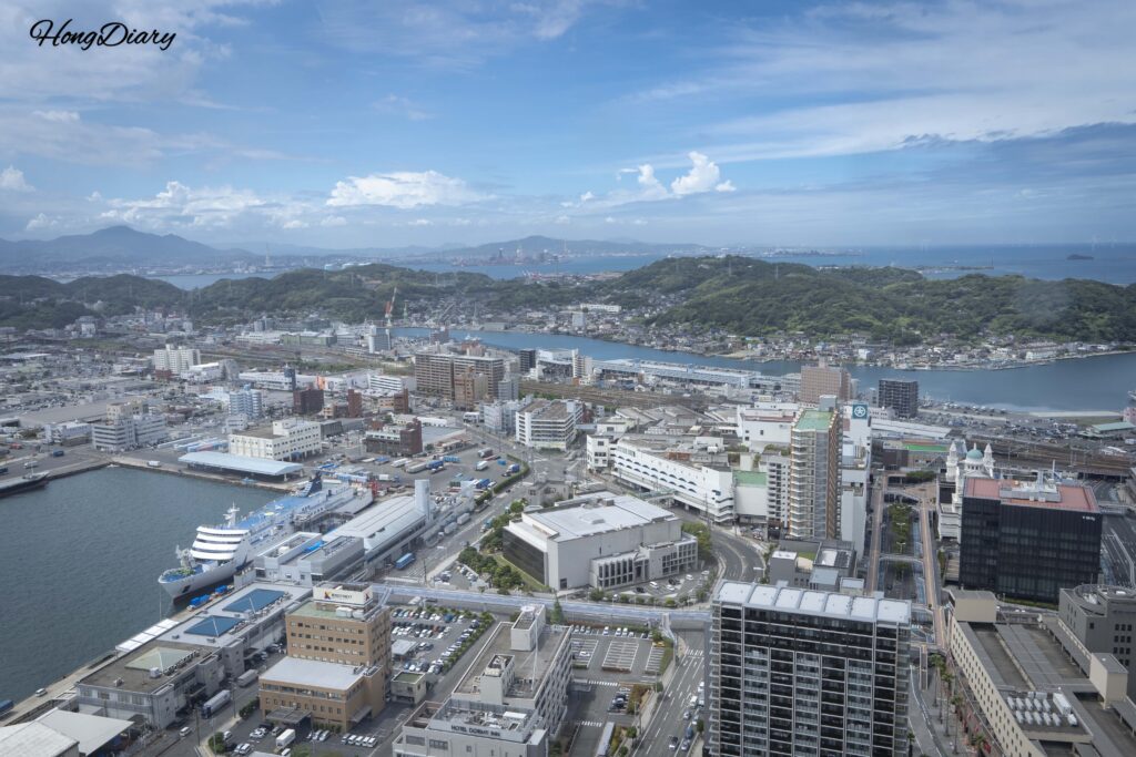 Vista hacia el oeste desde Kaikyo Yume Tower mostrando el ferry Kampu atracado en la Terminal Internacional del Puerto de Shimonoseki, la estación de Shimonoseki, vías de tren y el sol de la tarde reflejado en el mar