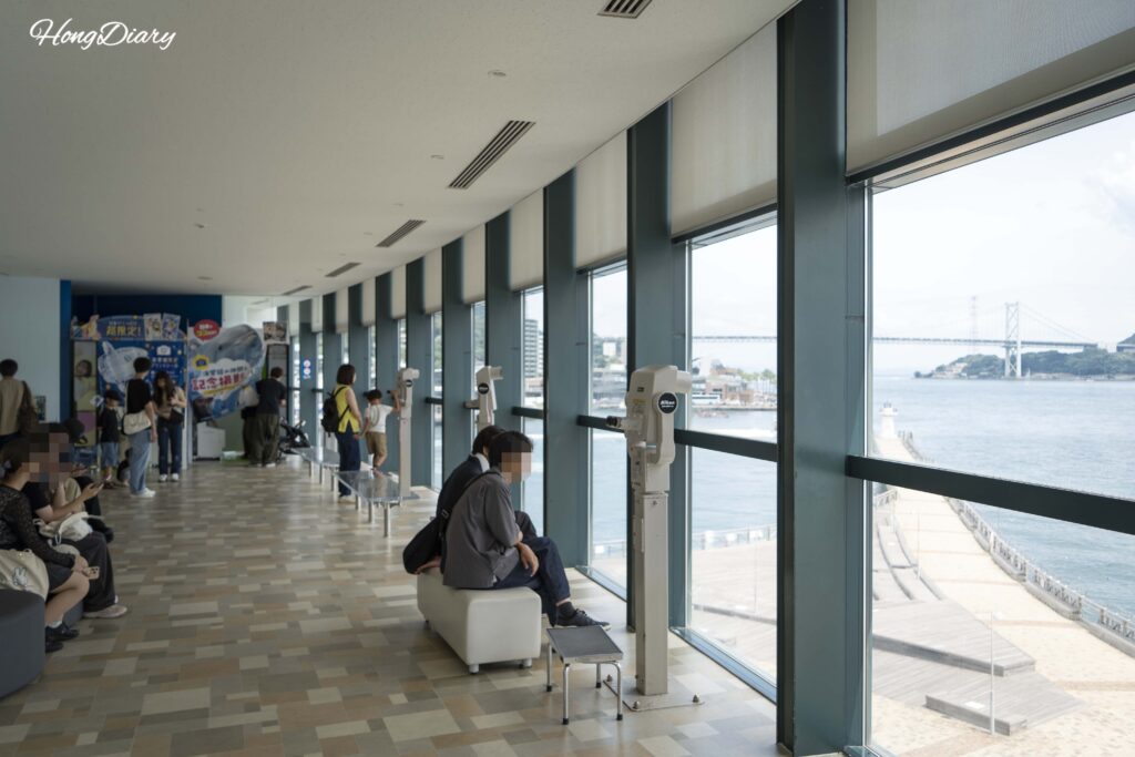 Visitors resting at a Kaikyokan Aquarium lounge area with a view of the Kanmon Bridge and strait through large windows