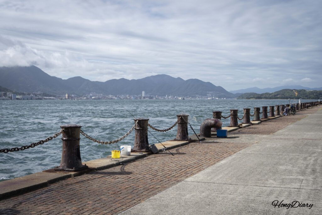 Rough waves crashing along the Kanmon Strait waterfront near RISONARE Shimonoseki, with a local fisherman casting a line on a windy day