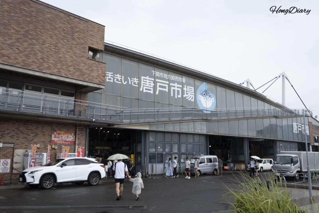 Entrada del Mercado Karato en Shimonoseki un día lluvioso, con visitantes sosteniendo paraguas y un niño con poncho de lluvia