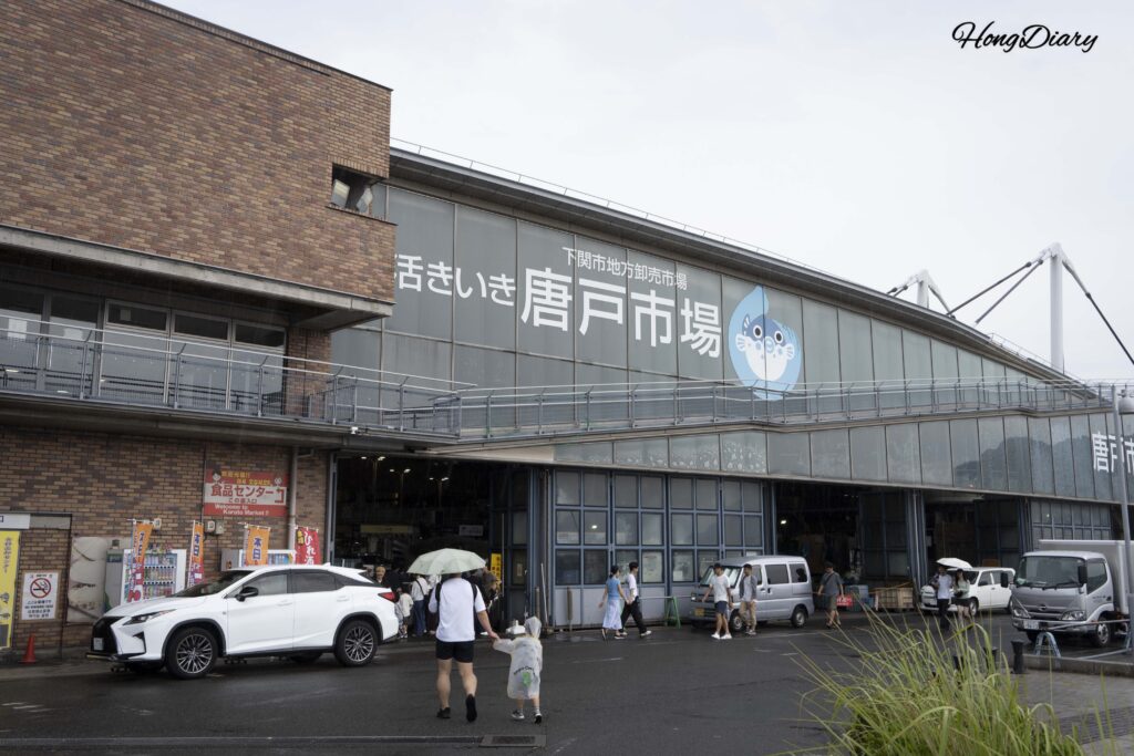 雨の日の唐戸市場入口、傘をさして歩く来場者とレインコートを着た子ども