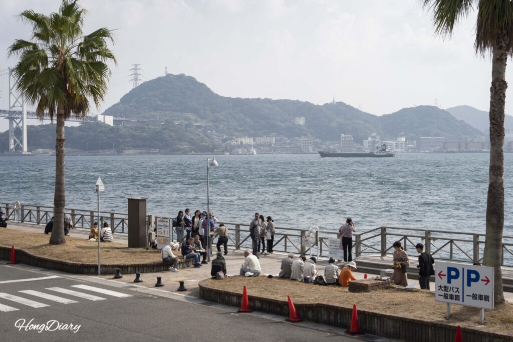 唐戸市場の屋外デッキで関門海峡と対岸の門司港を眺めながら寿司を食べる来場者と海峡を通過する貨物船