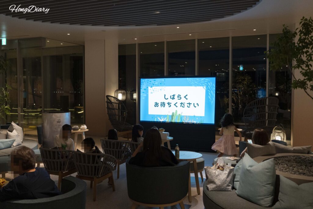 Guests watching the nightly Kaikyokan pufferfish lecture in the lobby of RISONARE Shimonoseki, with video content displayed on screen
