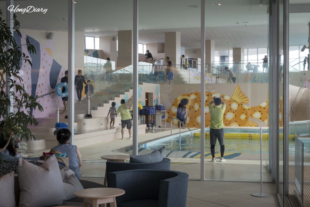 Indoor Fugu Pool at RISONARE Shimonoseki seen through the transparent glass wall from the lobby, with guests swimming
