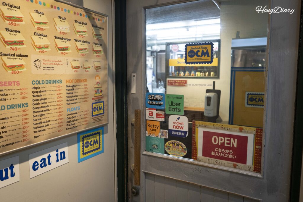 Second-floor entrance of Sandwich Factory OCM with a bilingual English and Japanese menu board displayed beside the door