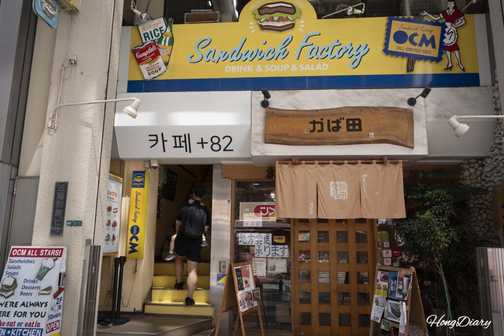 Fachada del edificio donde se encuentra Sandwich Factory OCM en Kokura, Kitakyushu, con una familia subiendo la escalera amarilla hacia el segundo piso