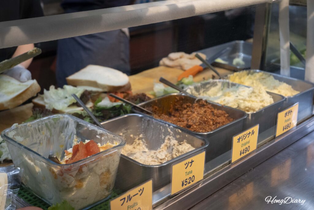 Close-up of fresh sandwich ingredients at OCM including tuna, fruit, original meat sauce, and potato salad with sandwiches being assembled in the background