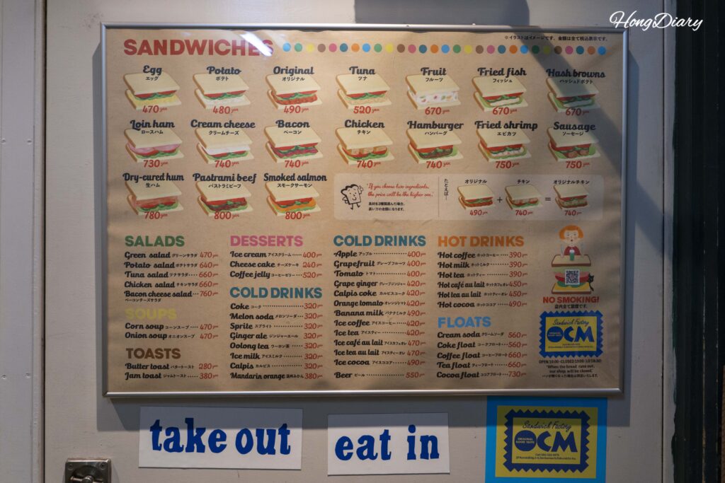 Full menu board at Sandwich Factory OCM in Kitakyushu showing all sandwich fillings, sides, drinks, and prices in English and Japanese with eat-in and take-out information