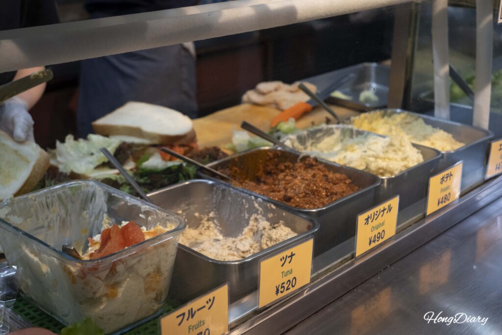 Primer plano de ingredientes frescos para sándwiches en OCM: atún, frutas, salsa de carne original y ensalada de papa, con sándwiches preparándose al fondo