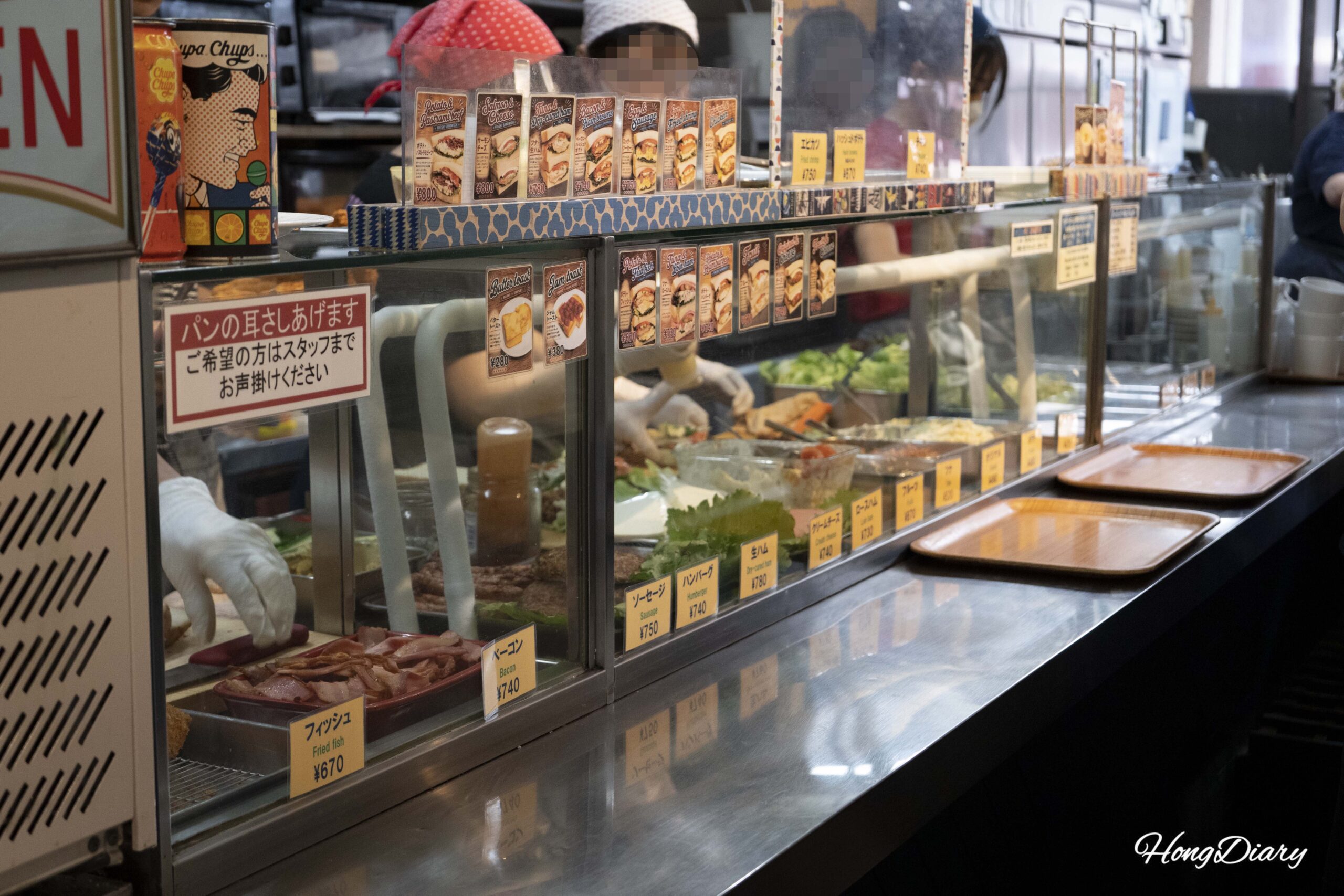 Sandwich ordering counter at OCM in Kokura showing recommended combo images with English and Japanese descriptions and fresh ingredients behind the glass