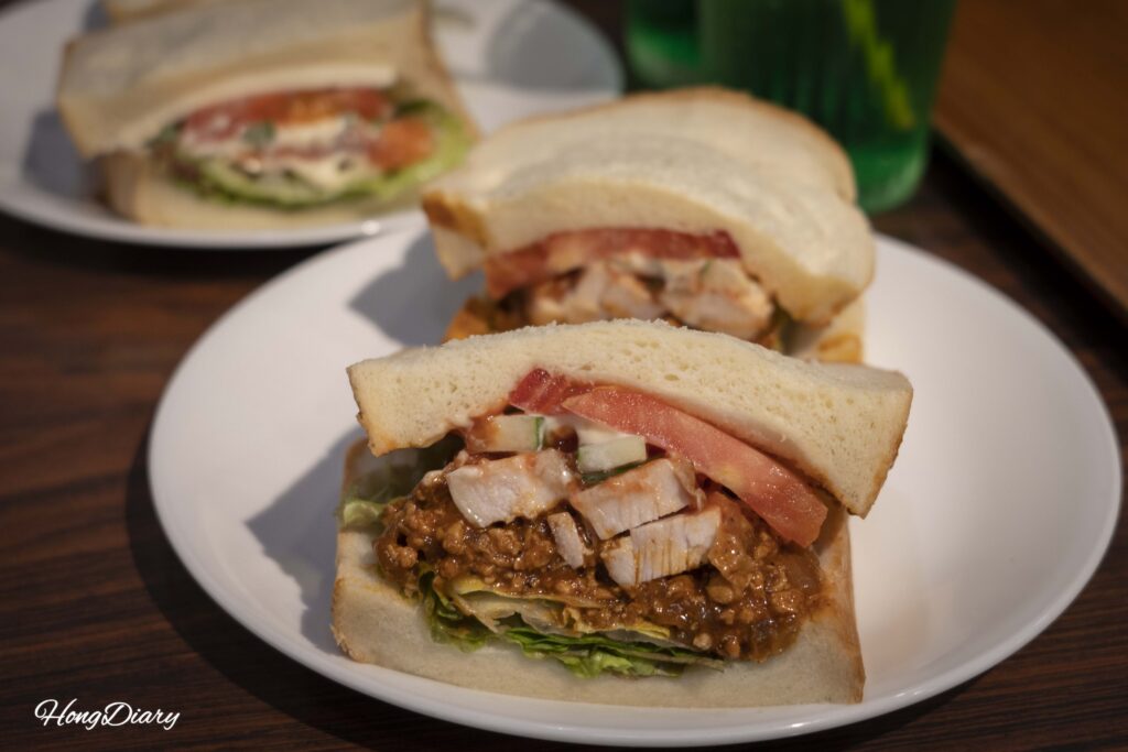 Primer plano del sándwich de Original y Pollo en OCM Kitakyushu, con rellenos generosos de salsa de carne de la casa y pollo de Kyushu entre pan blanco suave