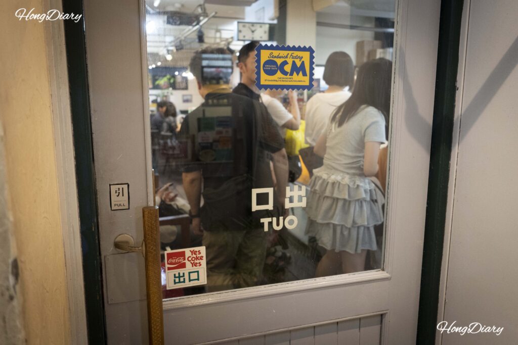 Vista desde la salida de Sandwich Factory OCM con clientes aún haciendo fila adentro y el sticker del logo de OCM pegado en la puerta de vidrio