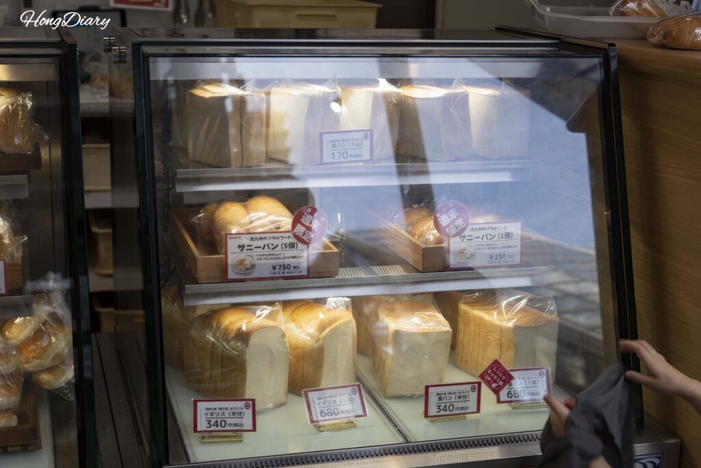 Bread display case at Shiroya Bakery in Kitakyushu showing various freshly baked items ranging from 150 to 750 yen, the bakery that supplies bread to Sandwich Factory OCM