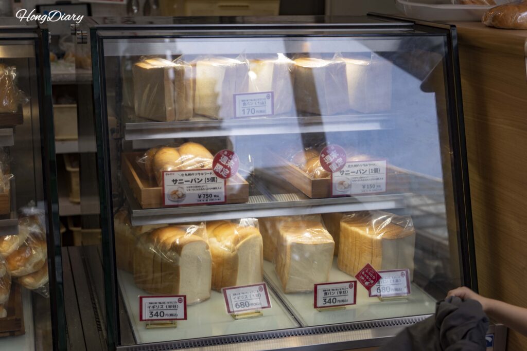 Vitrina de Shiroya Bakery en Kitakyushu con diversos panes recién horneados de ¥150 a ¥750, la panadería que provee el pan a Sandwich Factory OCM
